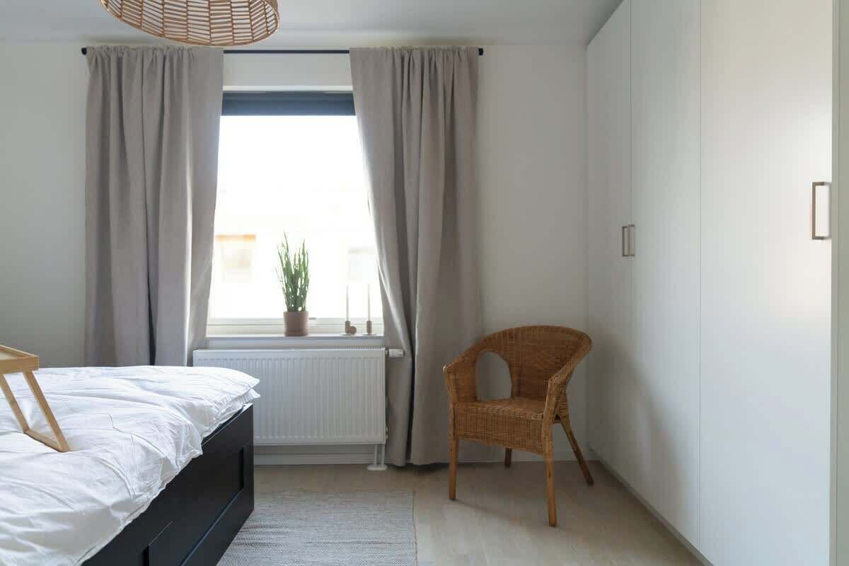 Un dormitorio con cama, una silla, un ventanal con cortinas y un radiador.