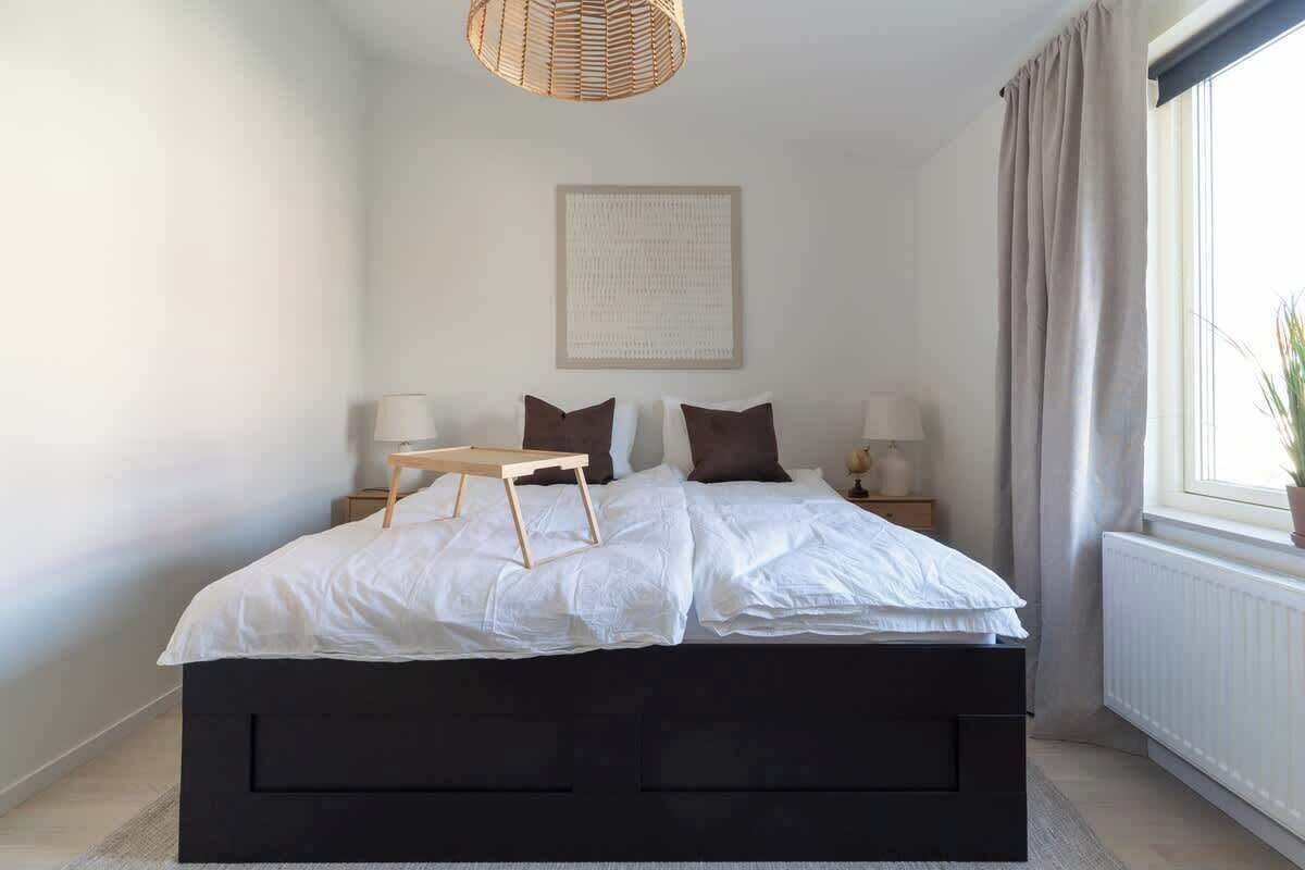 Un dormitorio con una cama grande, lámparas de noche, una mesita y una ventana con cortinas.
