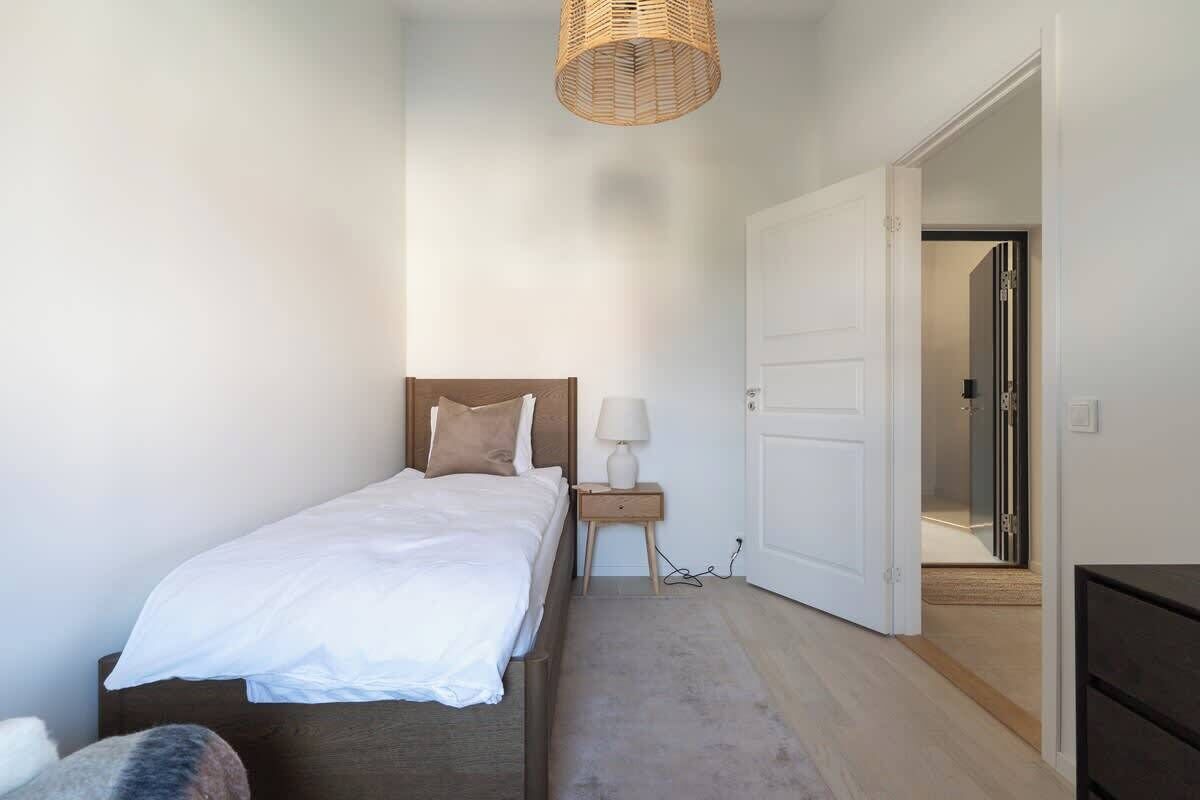 Een slaapkamer met een bed, nachtkastje, lamp en een deur naar een andere kamer.