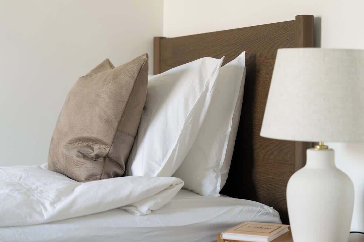 A neatly made bed with white and beige pillows, a wooden headboard, and a white bedside table with a lamp.