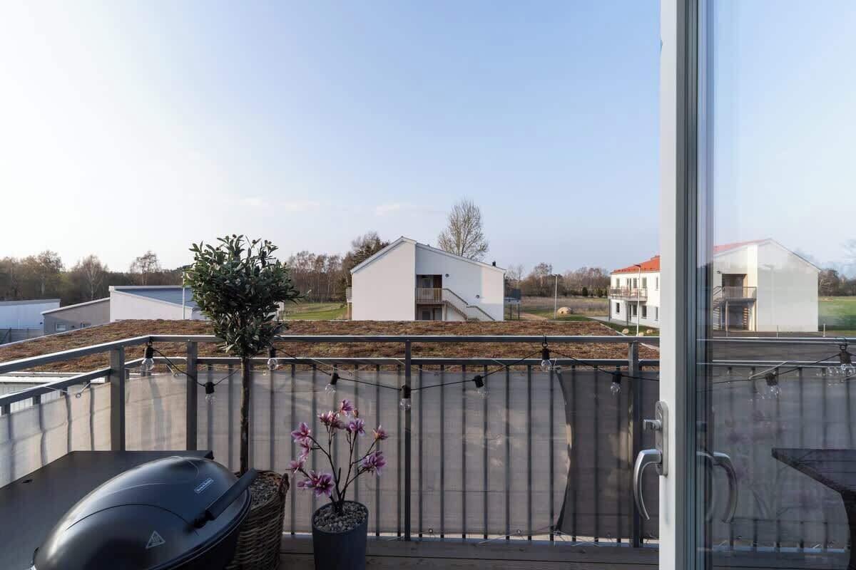 En balkon med grill, en potteplante og udsigt til en moderne bygning med rødt tag.