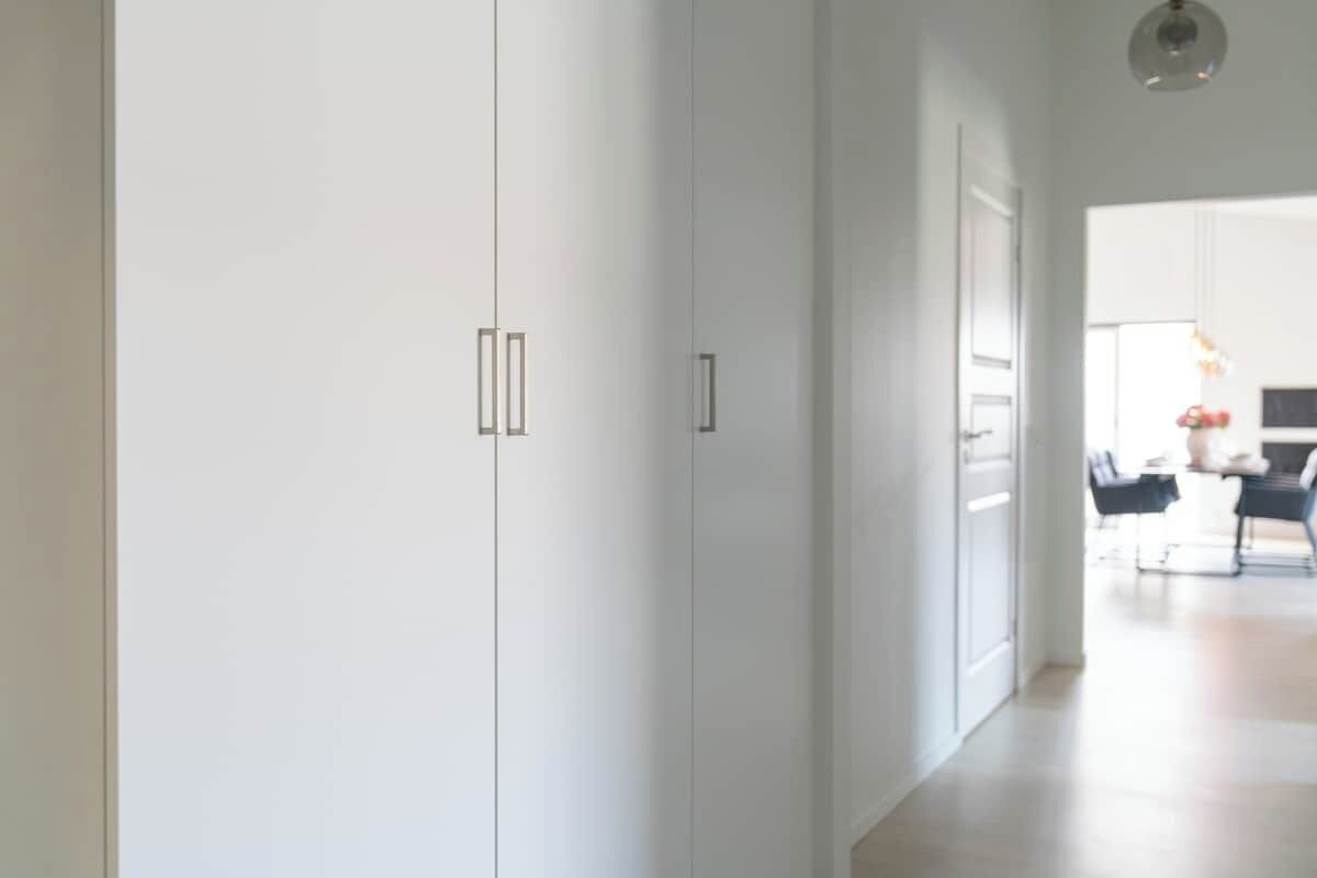 Ein moderner, minimalistischer Flur mit weißen Wänden und einem hellen Boden. Eine Tür rechts führt in einen Küchenbereich mit Esstisch und Stühlen.