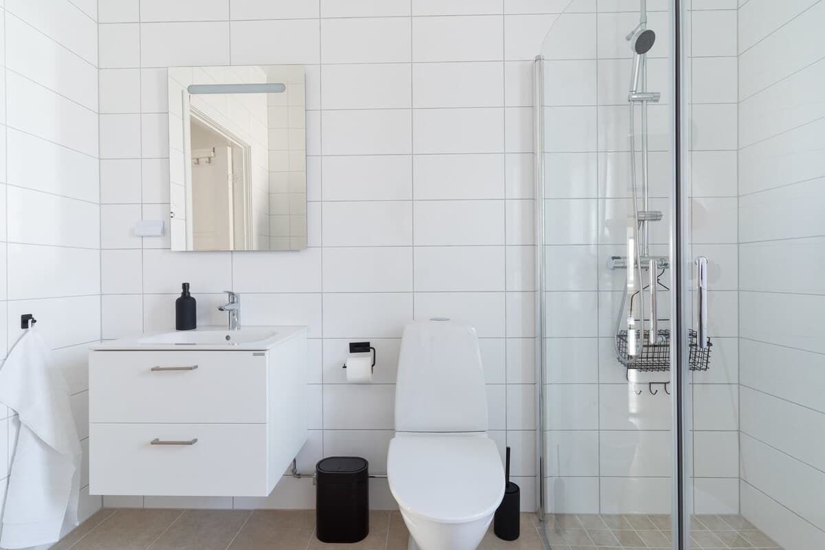A modern bathroom with white tiles, a white sink, a toilet, a mirror, and a glass-enclosed shower area.