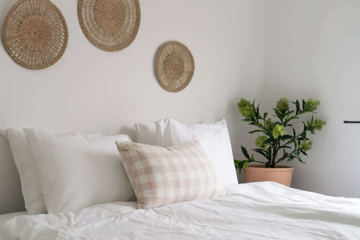 Een net opgemaakt bed met witte lakens en een geruit kussen, een potplant met groene bladeren en drie geweven wanddecoraties.
