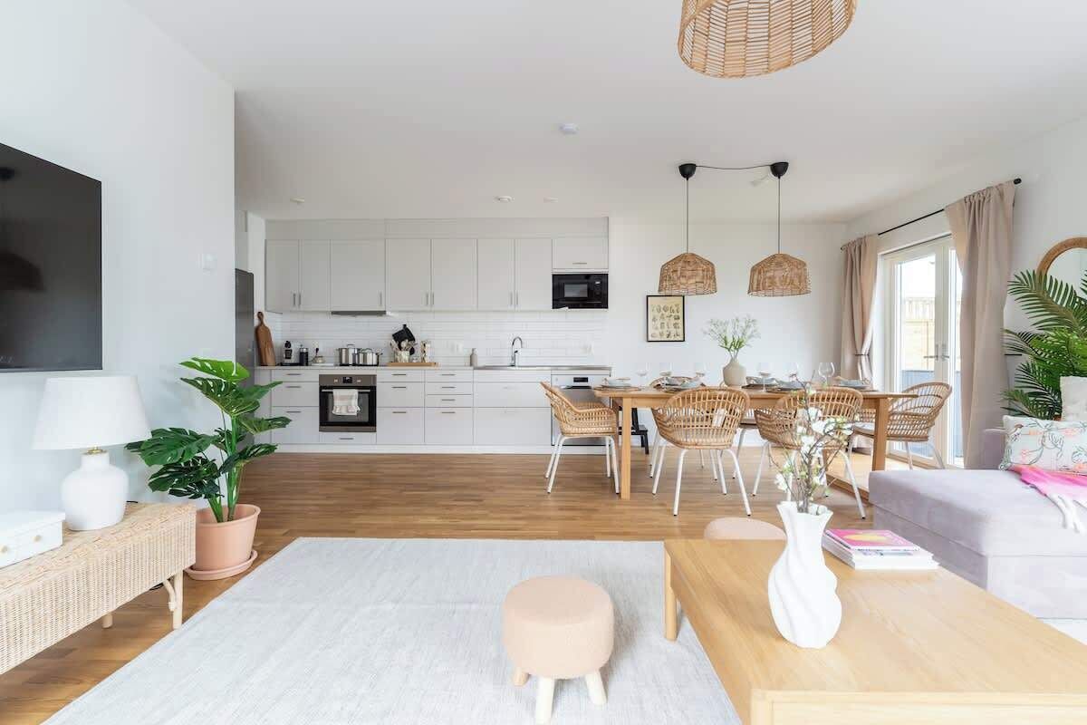 Una cocina moderna con armarios blancos, un comedor con sillas de mimbre y una sala de estar con sofá y mesa de centro.