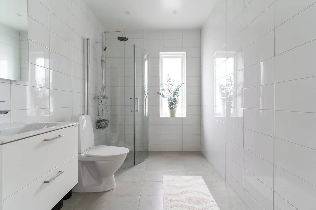 Un baño moderno con azulejos blancos, ducha con cerramiento de vidrio, inodoro y ventana que proporciona luz natural.