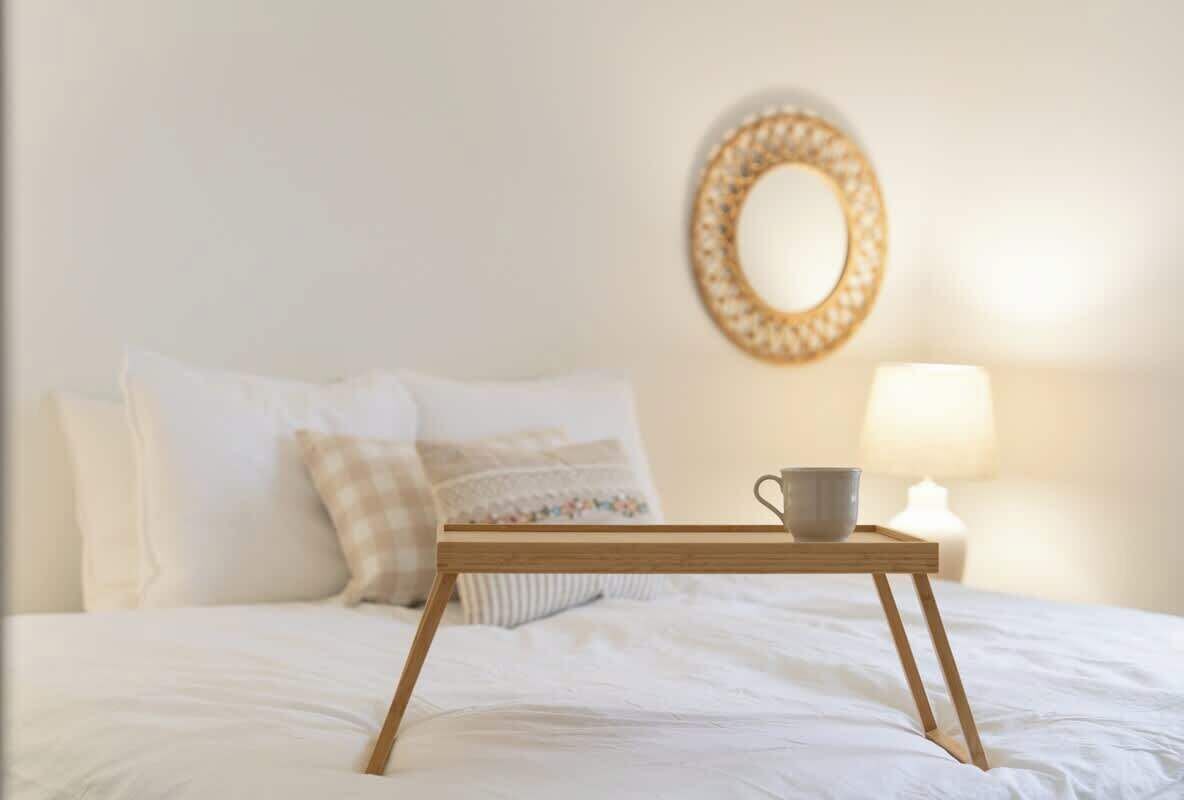 Uma cama com lençóis brancos, uma mesinha de cabeceira com uma xícara e um abajur, e um espelho decorativo na parede.
