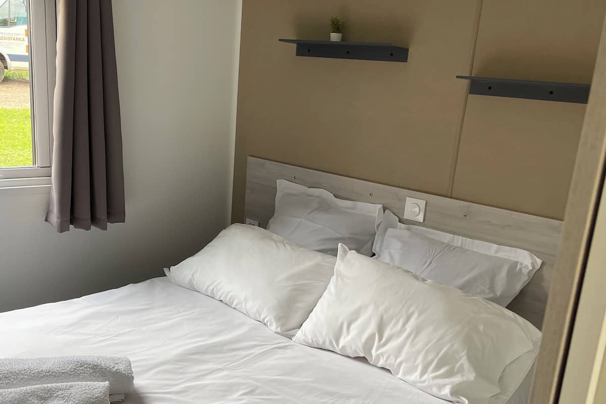 Un lit bien fait, avec du linge de lit blanc et une serviette pliée, une fenêtre avec des rideaux et une étagère fixée au mur, sur laquelle se trouve une petite plante.