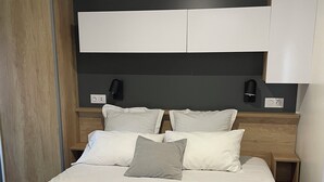Een net opgemaakt bed met witte lakens en een opgerolde handdoek, een houten hoofdbord, bedlampjes en een donkere muur.