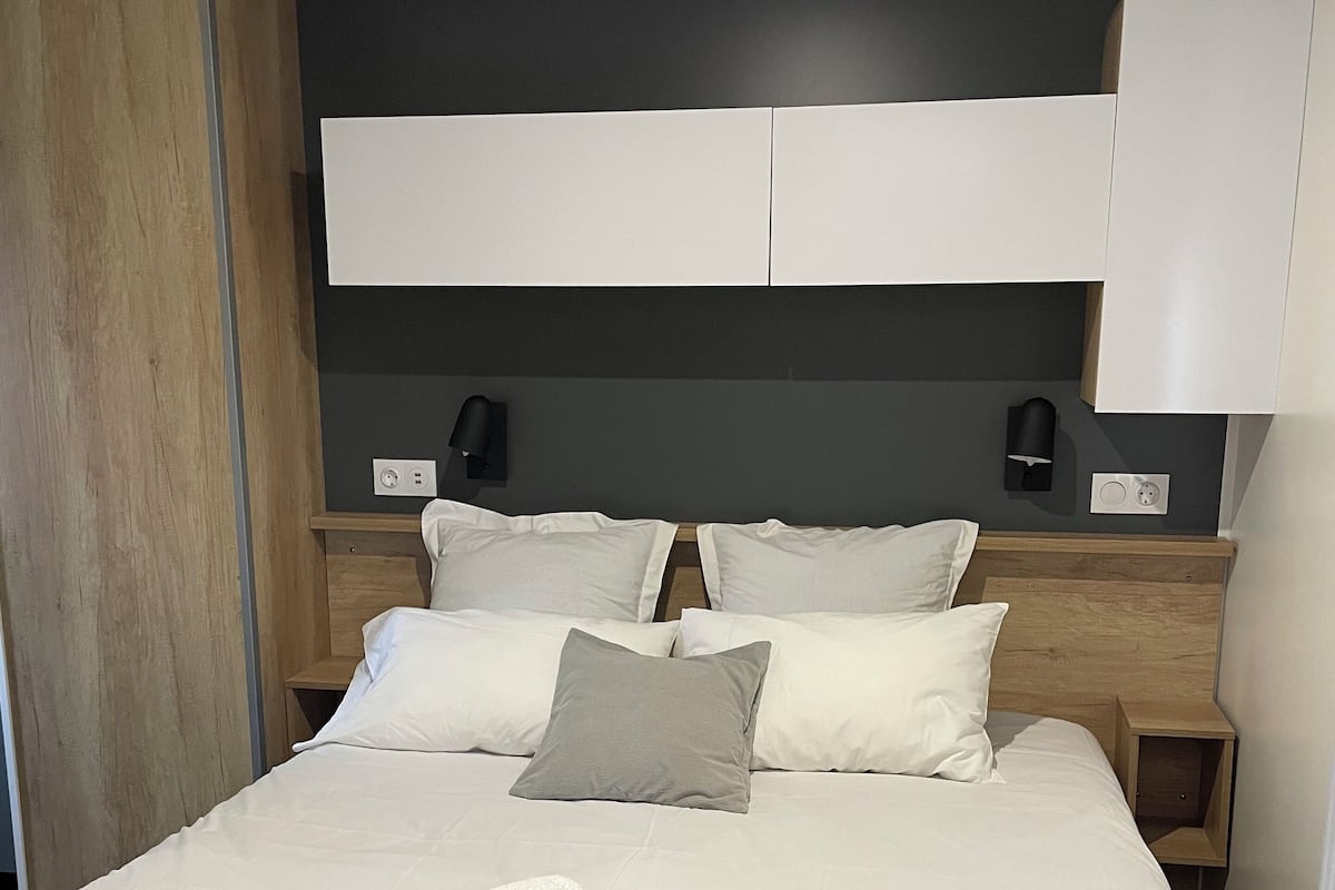 Un lit bien fait, avec du linge de lit blanc et une serviette enroulée, une tête de lit en bois, des lampes de chevet et un mur de couleur sombre.
