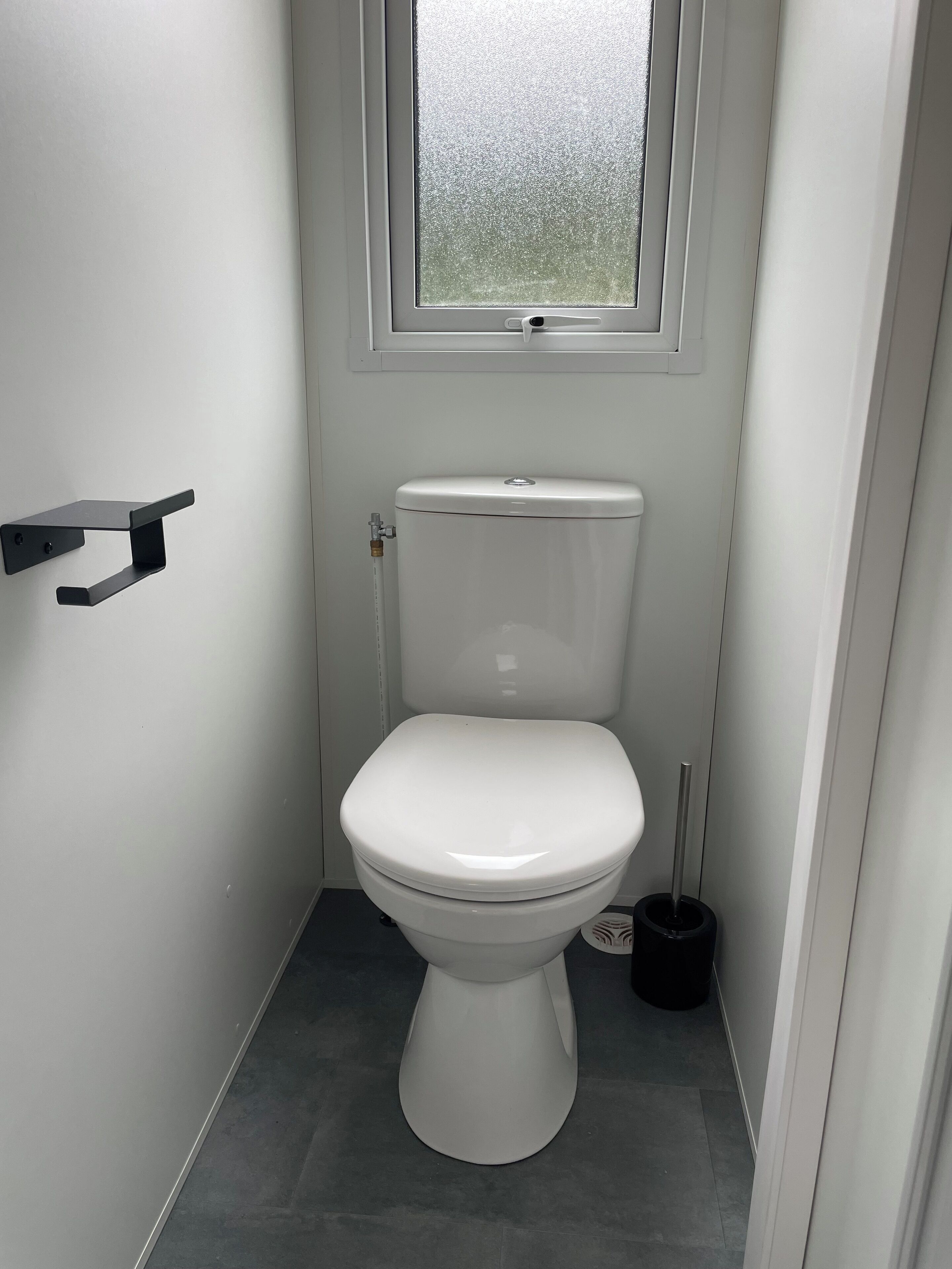 A small, white bathroom with a toilet, a window, and a black cleaning brush.