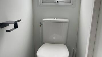 A small, white bathroom with a toilet, a window, and a black cleaning brush.
