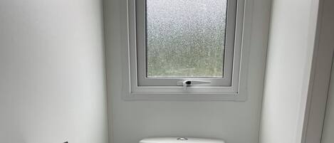 Un baño pequeño y blanco con inodoro, ventana y un cepillo de limpieza negro.