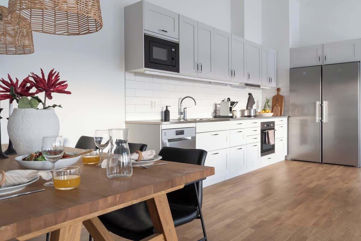 Una cocina moderna con armarios blancos, un refrigerador de acero inoxidable y un comedor de madera con sillas.