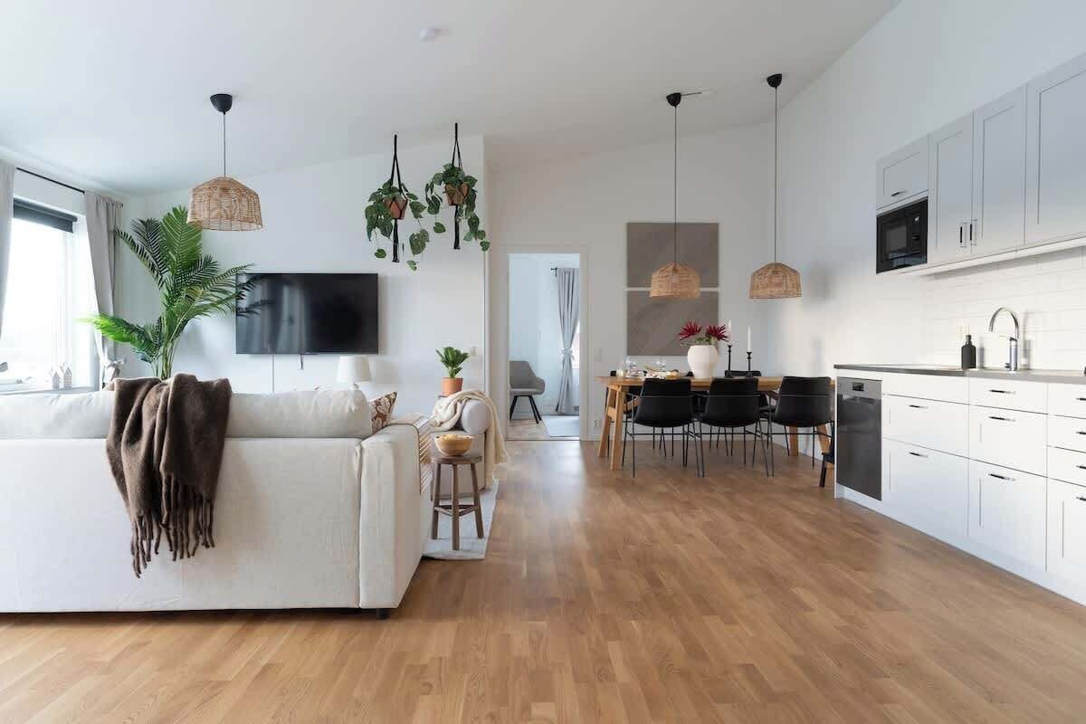 En rummelig opholdsstue med sofa, spisebord med stole, et køkken med hvide skabe og hængende planter.