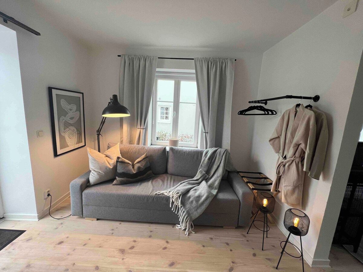 A cozy living room with a grey sofa, a standing lamp, a framed picture on the wall, and a window with curtains.
