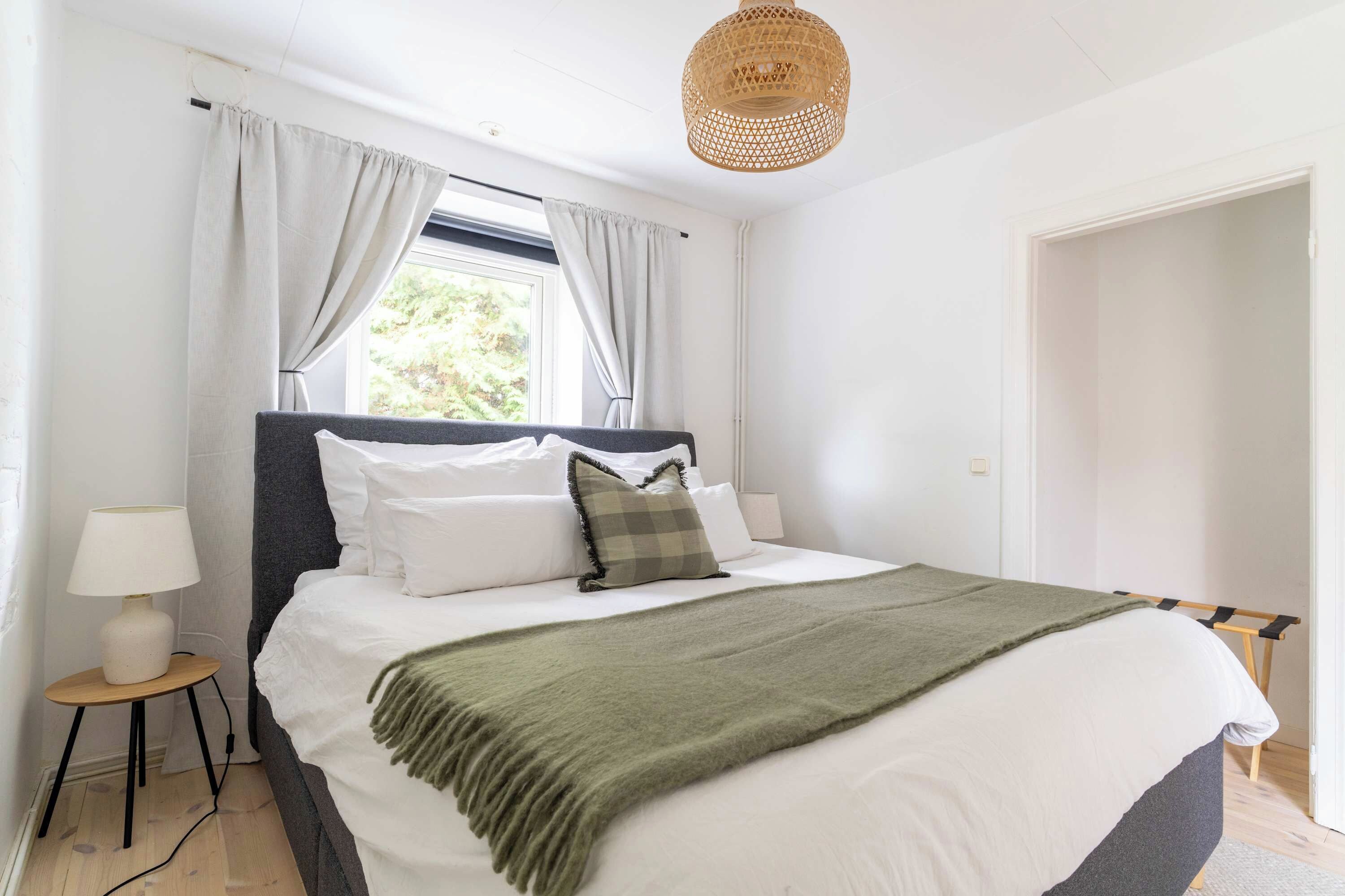A neatly made bed with white linens and a green throw, a bedside lamp, and a window with curtains.