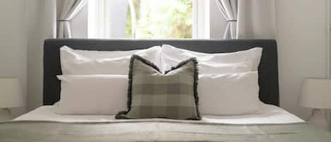 A neatly made bed with white linens and a checkered pillow, positioned in front of a window with curtains.