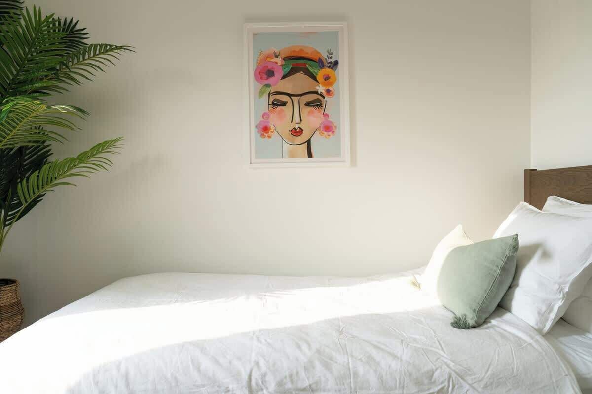 A bedroom with a bed, pillows, a framed artwork, and a potted plant.