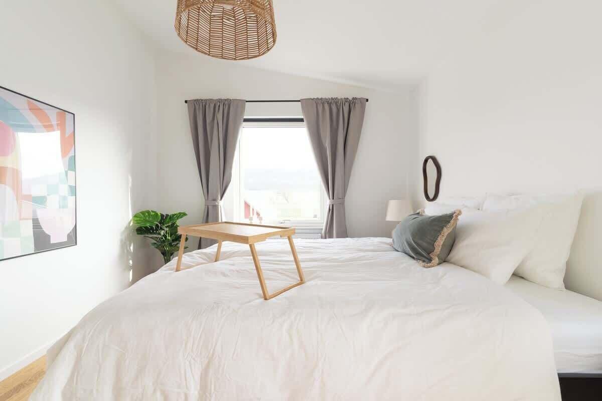 A bedroom with a large bed, a small wooden table, a potted plant, and a view of the outdoors through a window with curtains.