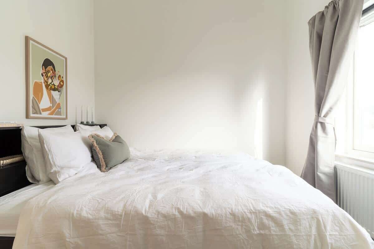 A neatly made bed with white linens and a few pillows, a bedside table with a lamp, and a framed artwork on the wall.