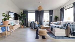 A modern living room with a sofa, coffee table, and TV. There are plants, curtains, and a large window.