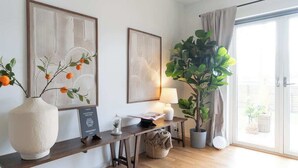 A modern living room with a wooden floor, a large white vase with orange flowers, two framed abstract artworks, a table with a lamp, a potted plant, and a sliding glass door leading to an outdoor area.
