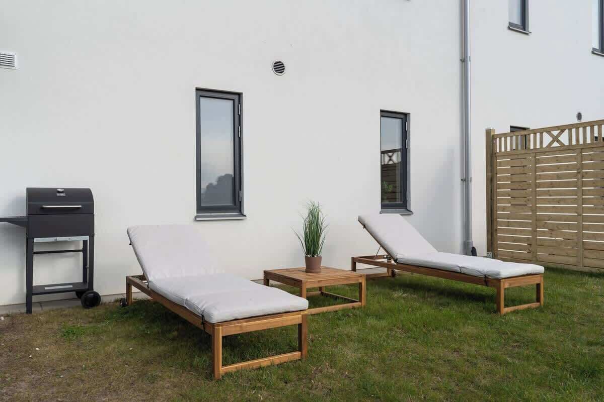 Två träloungestolar med kuddar, ett litet bord och en krukväxt i en trädgård med en vit vägg och ett trästaket.