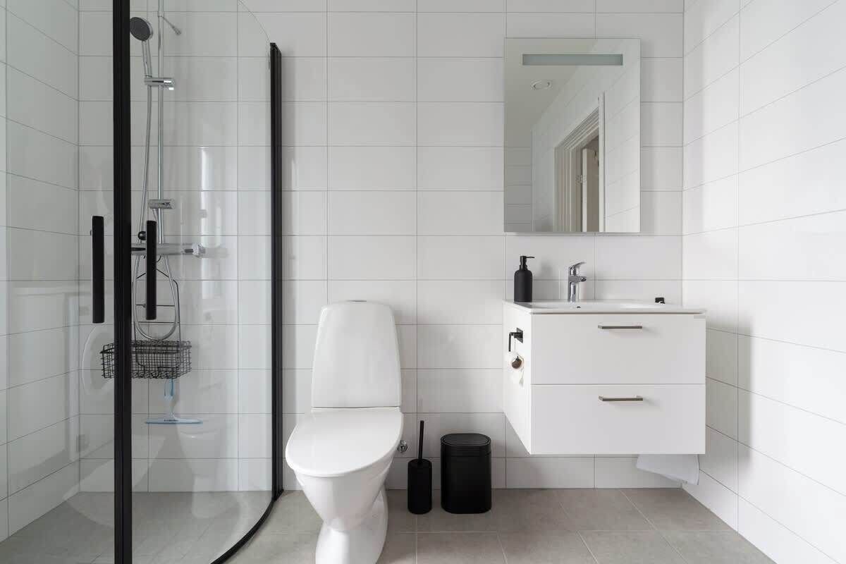 Un baño moderno con cabina de ducha de vidrio, inodoro blanco, lavamanos con espejo y un cubo de basura.