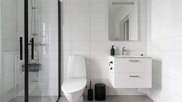 Un baño moderno con cabina de ducha de vidrio, inodoro blanco, lavamanos con espejo y un cubo de basura.