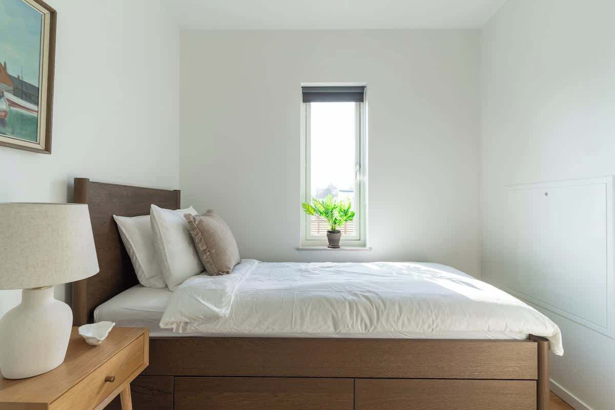 A bedroom with a bed, a bedside table with a lamp, a window with a plant, and a painting on the wall.