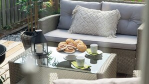 A wicker outdoor seating area with a cushioned sofa, a glass table with pastries and a candle, and a wooden deck. - Fully Equipped Apartment Close to Malmo Copenhagen (Vellinge)