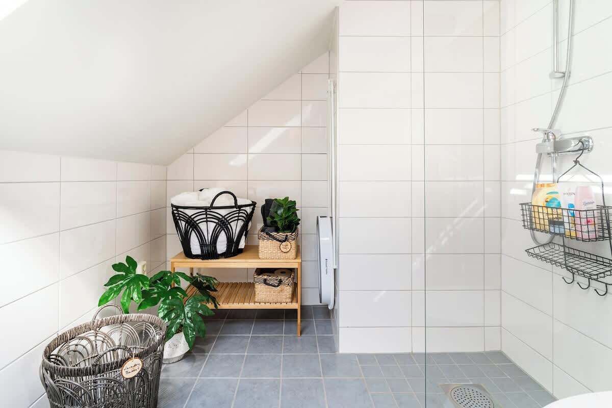 A small, modern bathroom with white tiles, a shower, a shelf with baskets, and a plant.