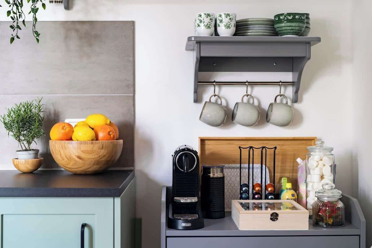 A kitchen with a wooden bowl of oranges, a coffee maker, and a shelf with cups and jars.