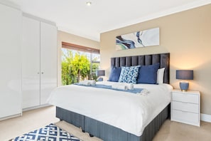 A bedroom with a large bed, a nightstand, a lamp, a wardrobe, and a view of greenery outside.
