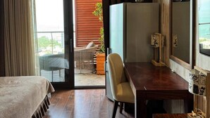A hotel room with a bed, a desk, a chair, a TV, and a balcony with a wicker chair and potted plant.