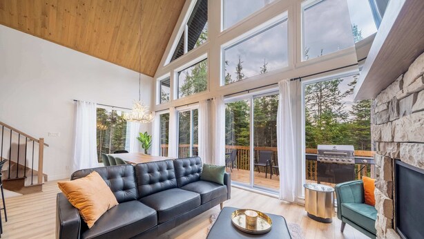 A modern living room with a black leather sofa, a stone fireplace, and large windows offering a view of the outdoors.