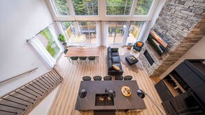 A spacious living area with a central table, seating, and a stone fireplace. Large windows provide views of the outdoors.