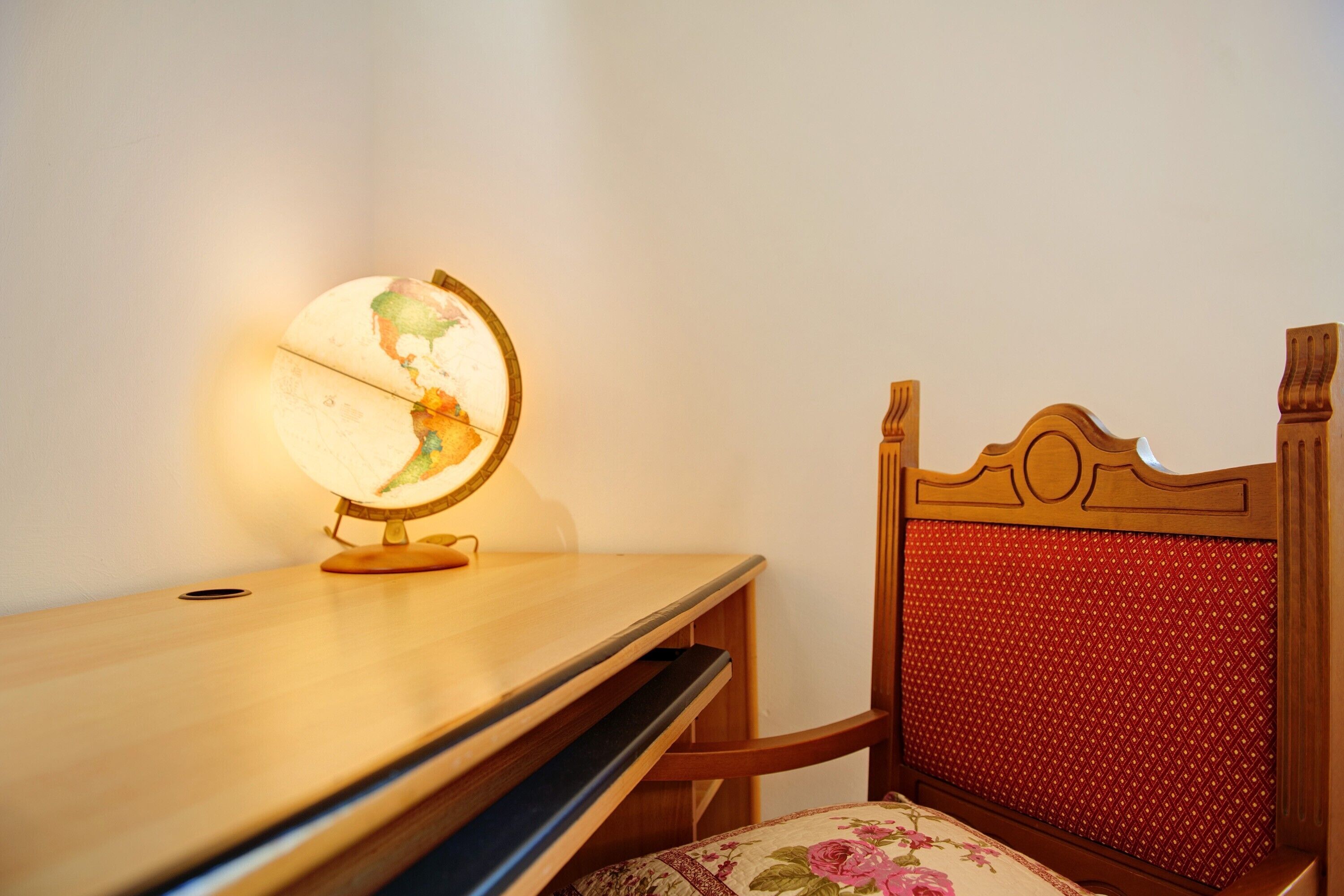 Ein Holztisch mit einer Globenlampe, ein Stuhl mit Sitzkissen in Blumenmuster und ein rotes, gepolstertes Kopfteil.