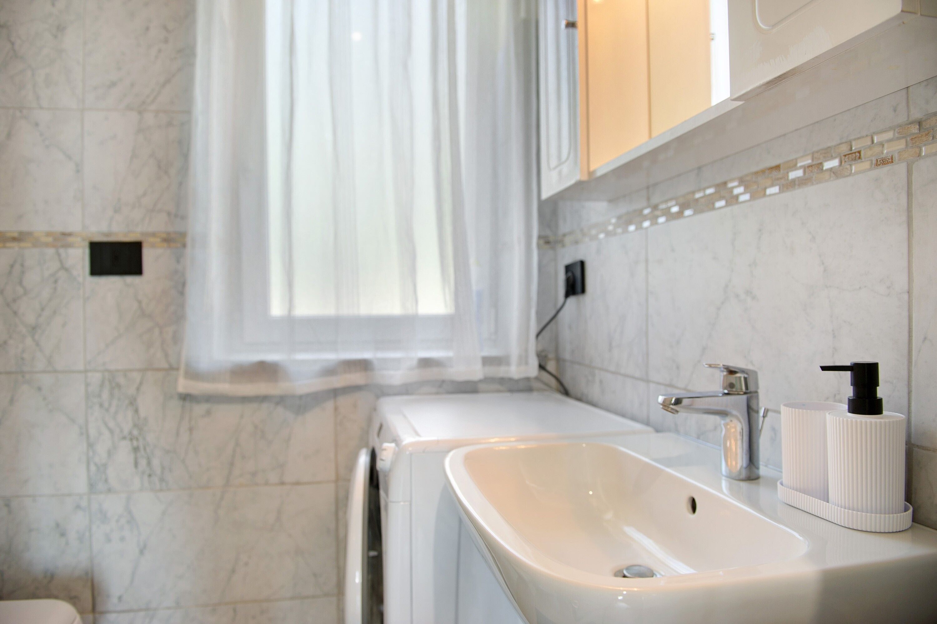 Une salle de bain avec un lavabo blanc, un miroir, un porte-serviettes et une fenêtre avec un rideau.