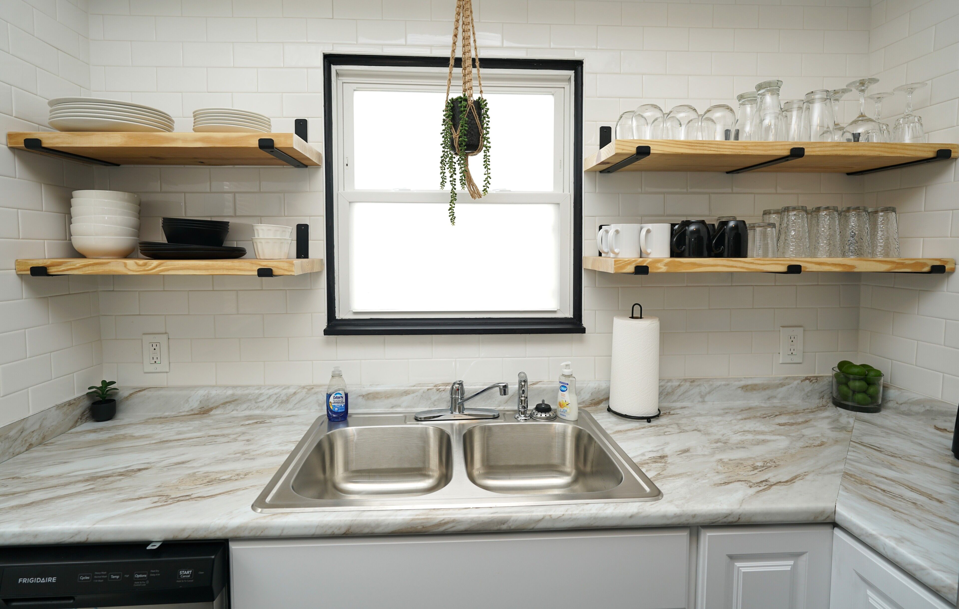 A kitchen with a double sink, white tiled backsplash, wooden shelves, and a hanging plant.