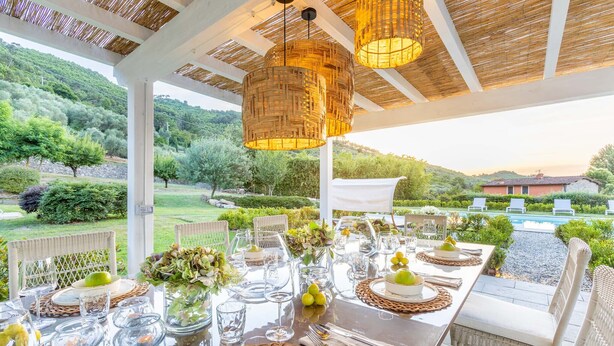 An outdoor dining area with wicker chairs, a table set for a meal, and a view of a pool and greenery.