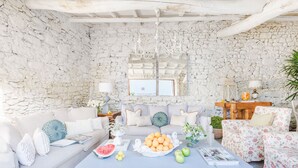 A cozy living room with a stone wall, a white sofa, a coffee table with fruits and magazines, and a floral armchair.