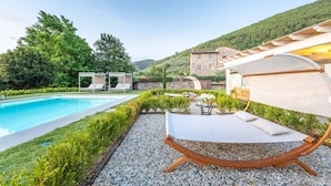 A poolside area with a lounge chair, a pergola, and a stone building in the background.