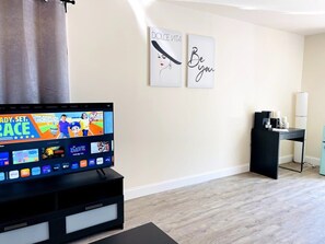 A living room with a flat-screen TV displaying a sports channel, a coffee table, a black desk with a lamp, and two framed wall art pieces. - #1 "The village" great Studio (North Myrtle Beach)