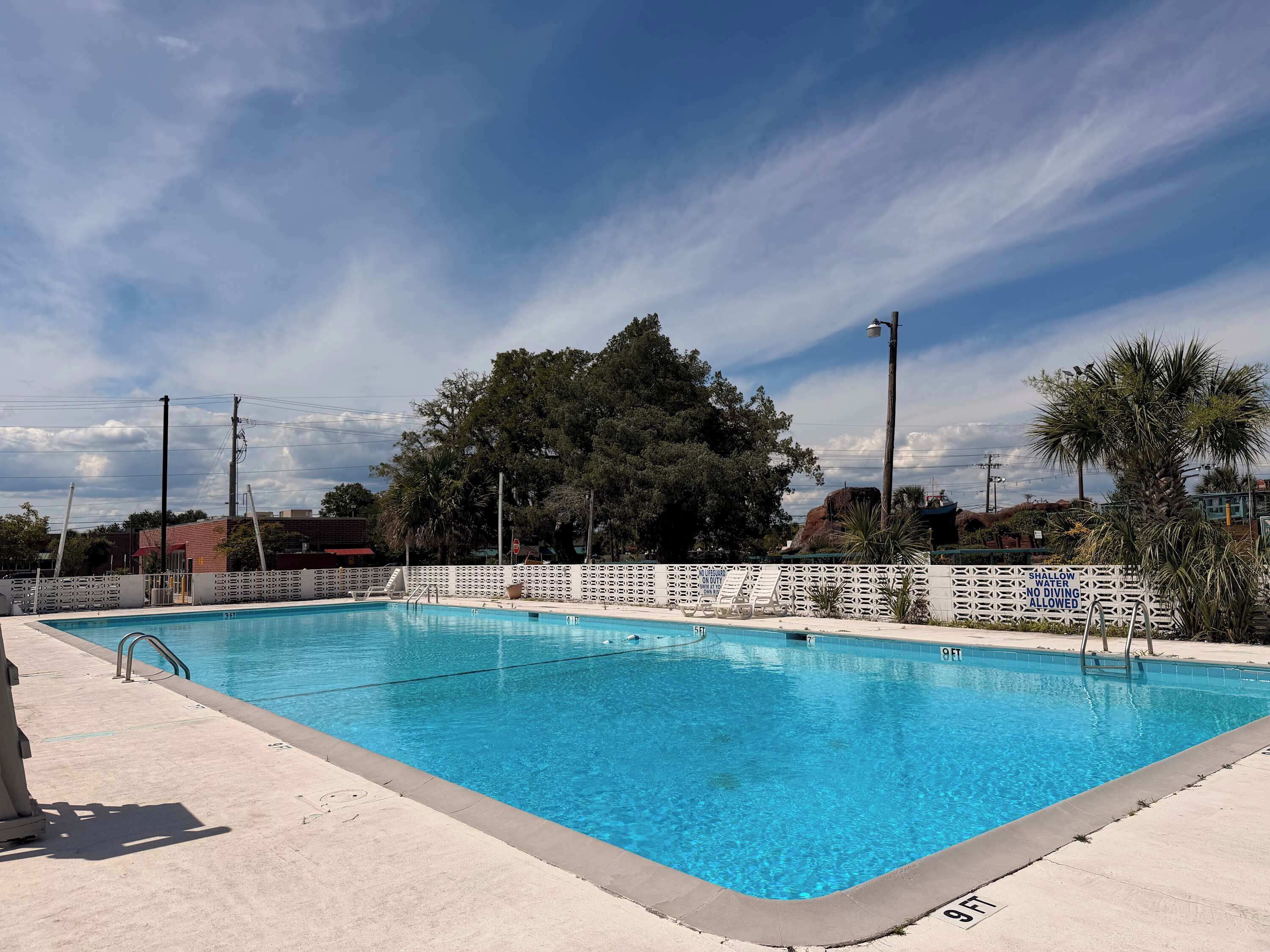 A clear blue swimming pool with a white fence, surrounded by trees and a cloudy sky.