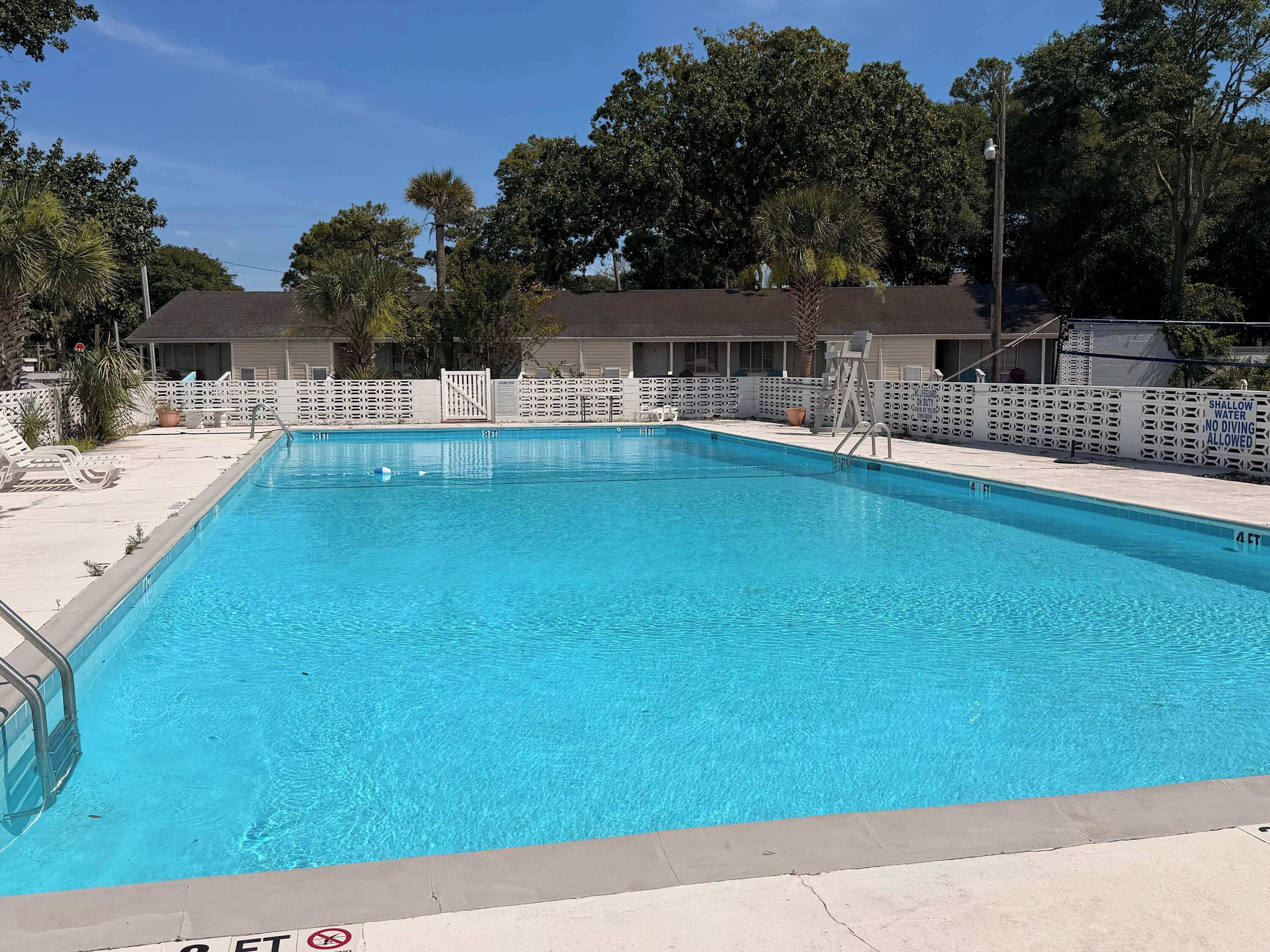 Ein rechteckiger Pool mit weißer Liegefläche, umgeben von einem weißen Zaun, und ein Gebäude mit überdachter Terrasse im Hintergrund.