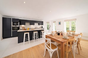 A modern kitchen with dark cabinets, a central island with stools, and a dining area with a wooden table and white chairs. - Spectacular Sea Views at Tors Park (Lynmouth)