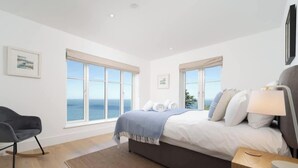 Une chambre à coucher comprenant un lit, une chaise, un bureau et deux fenêtres donnant sur la mer.