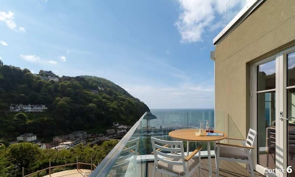 A balcony with a glass railing overlooking a coastal view, a round wooden table, and white chairs. - Spectacular Sea Views at Tors Park (Lynmouth)